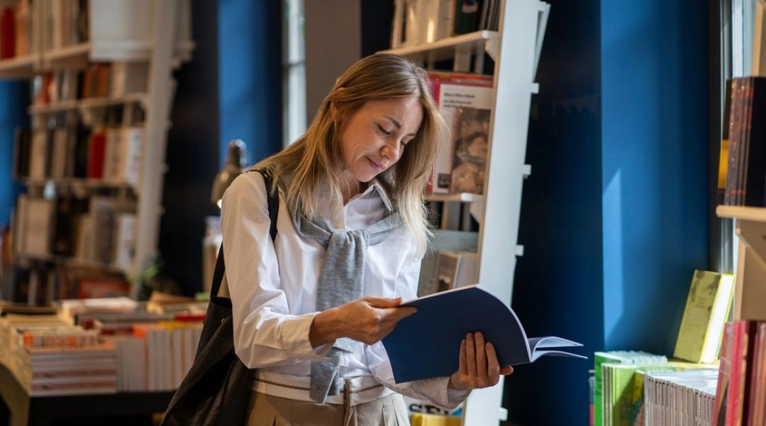 Kobieta przegląda książkę artystyczną w księgarni lub przestrzeni wystawienniczej z półkami pełnymi publikacji.
