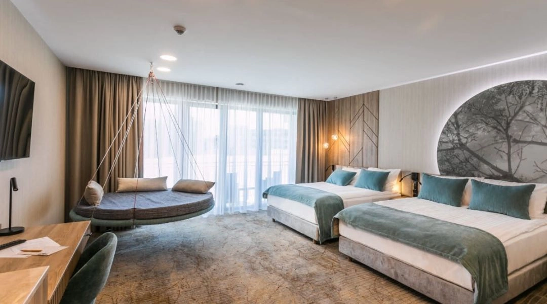 Przestronny hotelowy pokój z dwoma dużymi łóżkami, turkusowymi narzutami i wiszącą leżanką przy oknie.