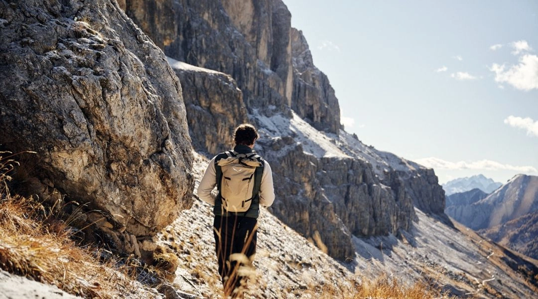 Turysta z plecakiem idzie wąską, skalistą ścieżką u podnóża stromych ścian Dolomitów.