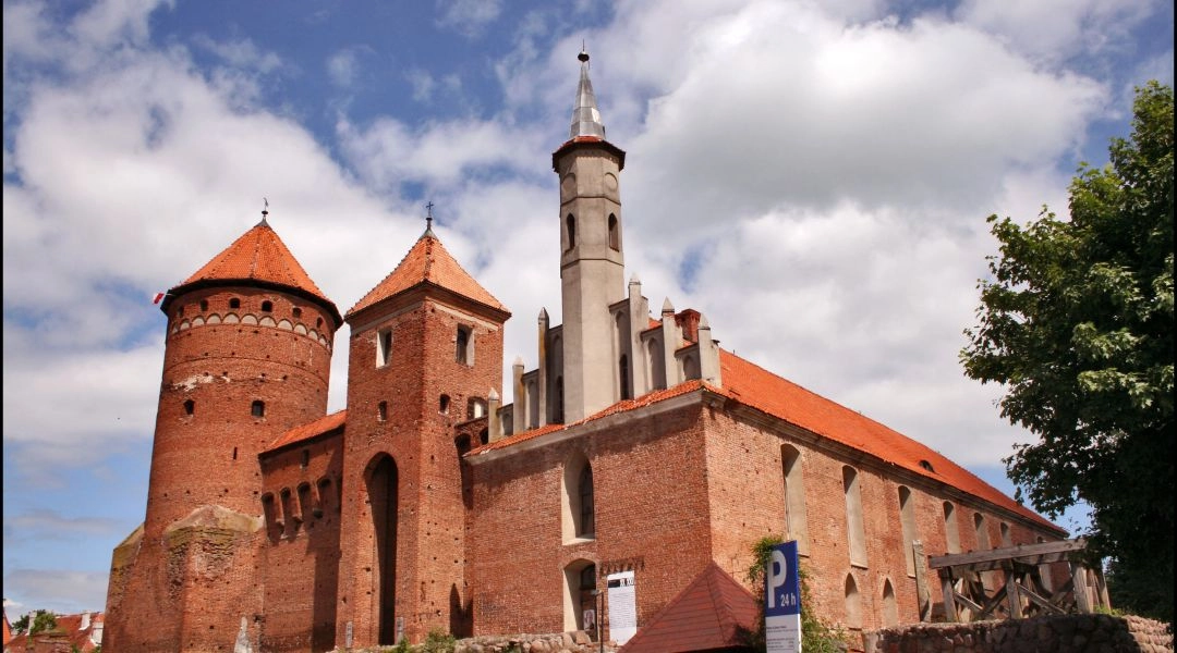 Zamek Biskupów Warmińskich w Reszlu z ceglaną wieżą i przylegającym kościołem pod błękitnym, częściowo zachmurzonym niebem.