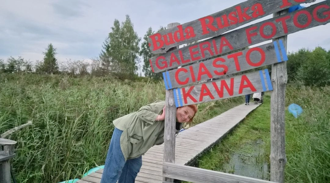 Drewniana tablica z napisem „Buda Ruska 16, Galeria Fotografii, Ciasto, Kawa” przy pomoście wśród wysokich trzcin i turystka wyglądająca zza tablicy.