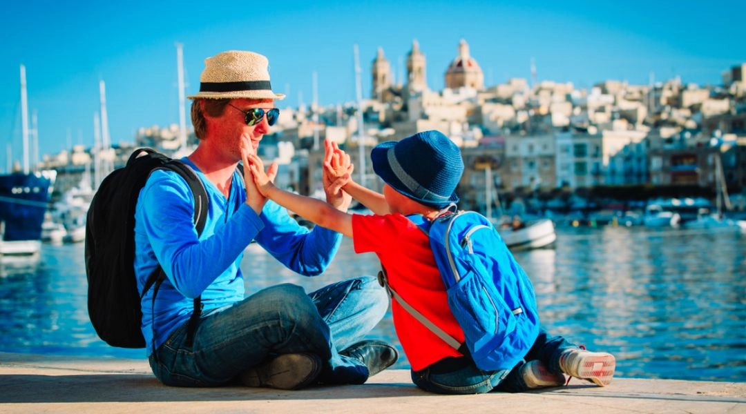 Rodzic z dzieckiem siedzący nad portem w maltańskim miasteczku, w tle zabudowa nadmorska i spokojna marina.