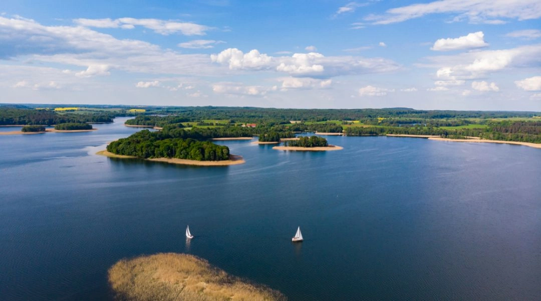 Mazury z lotu ptaka, żaglówki na jeziorze i zielone wyspy wśród wody.