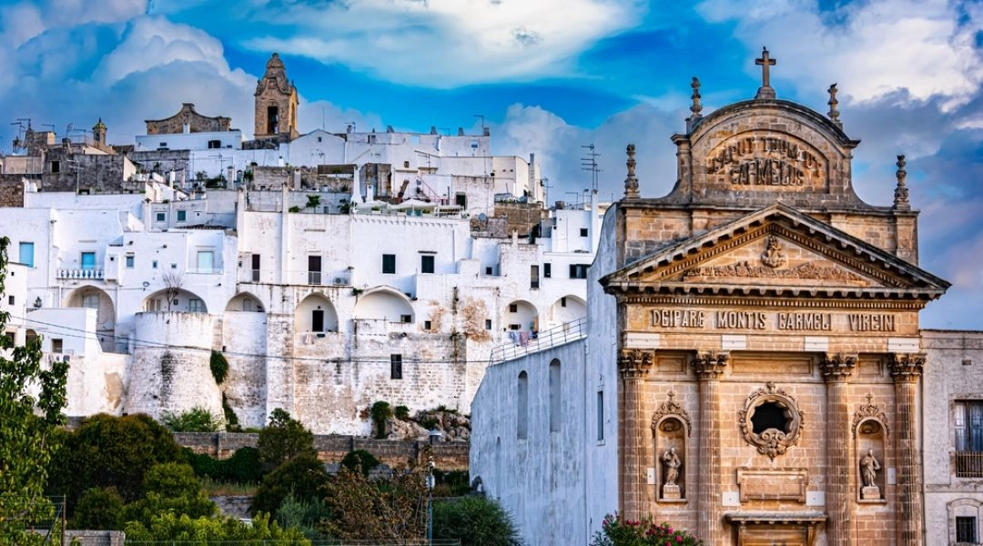 Białe domy Ostuni na wzgórzu oraz kamienna fasada kościoła z krzyżem na pierwszym planie.