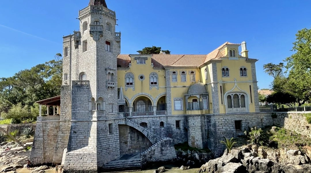 Pałacowa willa nad Atlantykiem w Cascais, z kamienną wieżą, żółtą fasadą i arkadowym przejściem nad wodą.