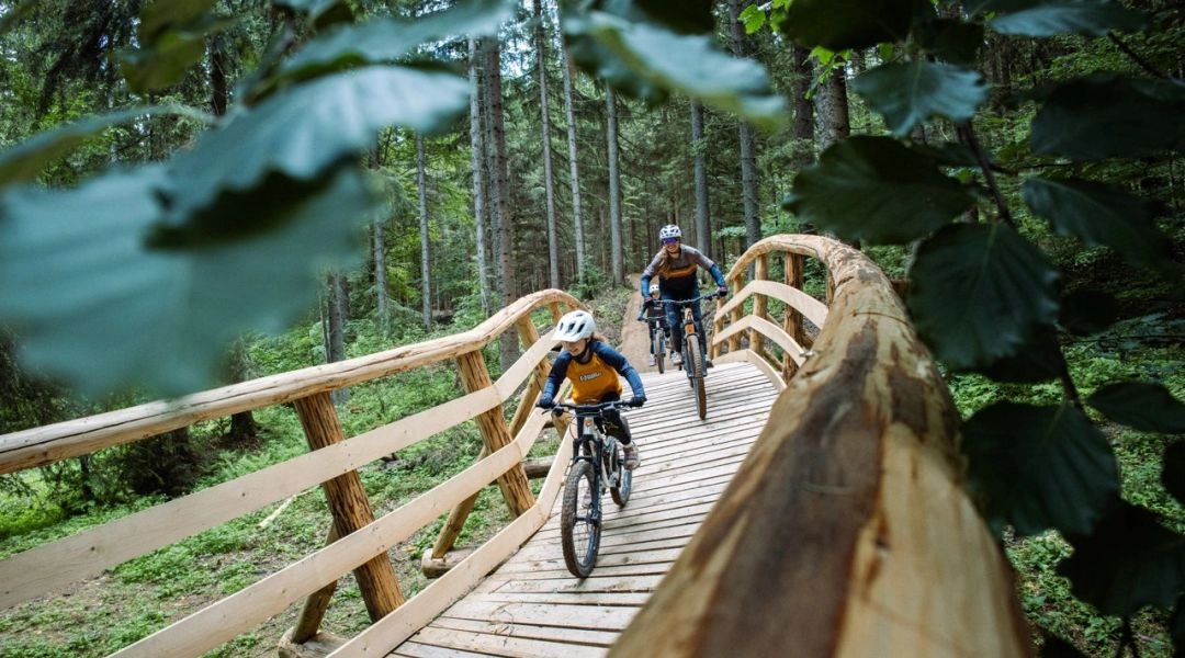 Grupa rowerzystów jedzie trasą w Donovalach na tle hoteli, lasów i górskich szczytów.