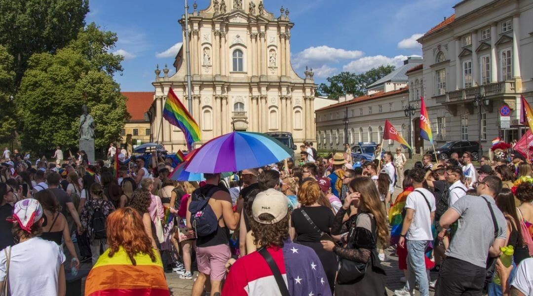 Uczestnicy Parady Równości w centrum Warszawy z tęczowymi flagami i kolorowym parasolem.
