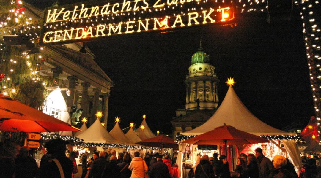Wejście na jarmark WeihnachtsZauber na Gendarmenmarkt w Berlinie z oświetlonymi stoiskami i kopułą katedry w tle.