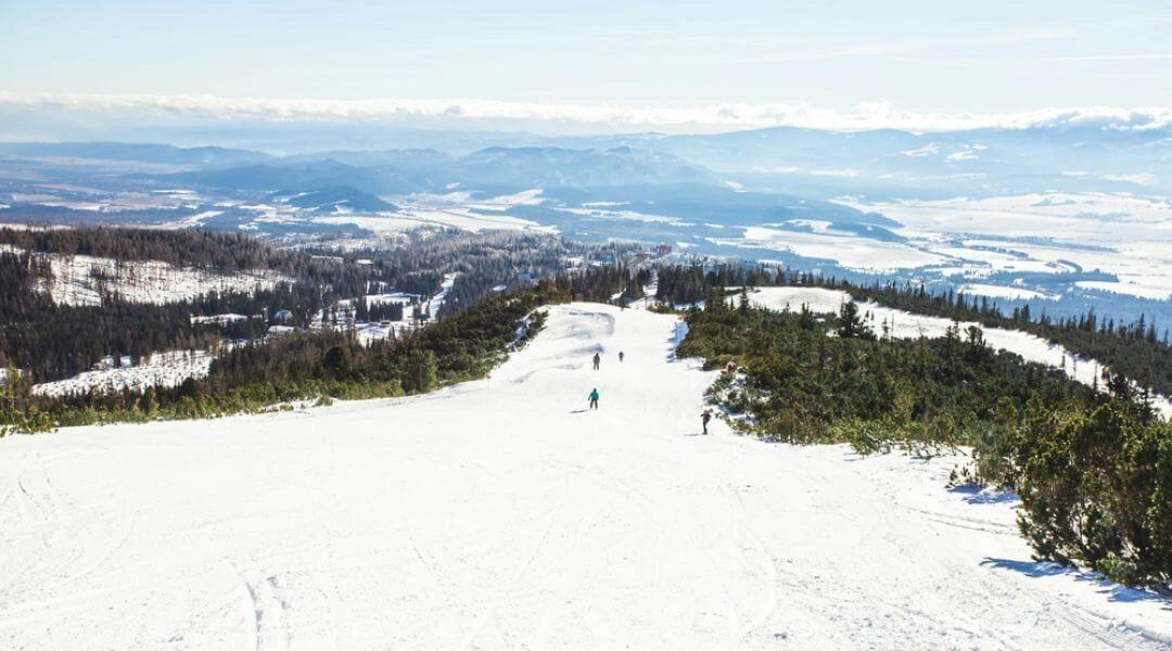 Szeroka trasa narciarska w rejonie Szczyrbskiego Plesa z widokiem na dolinę i ośnieżone szczyty w oddali