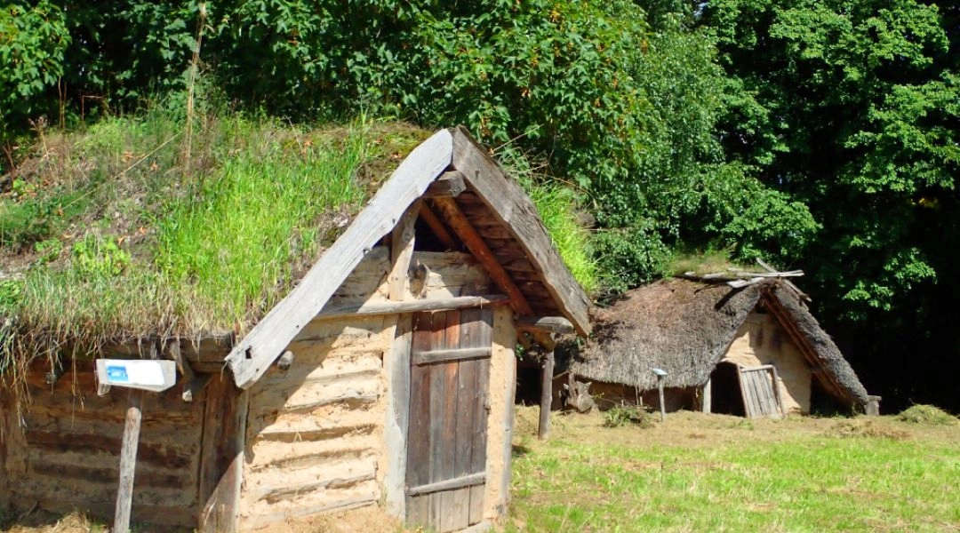Rekonstrukcje niewielkich drewnianych chat częściowo zagłębionych w ziemi, otoczonych zielenią.