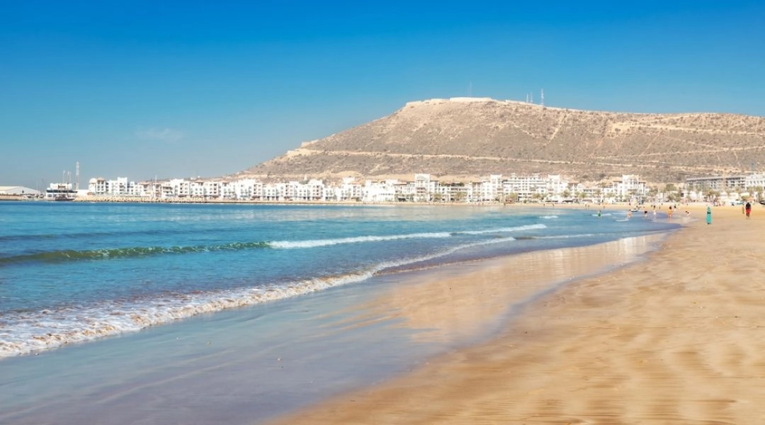 Szeroka piaszczysta plaża w Agadirze z widokiem na wzgórze Agadir Oufella i białą zabudowę miasta.