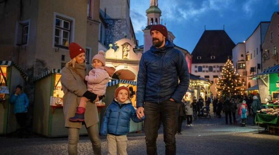 Rodzina z dwójką dzieci spaceruje wieczorem po tyrolskim jarmarku adwentowym w otoczeniu drewnianych straganów i świątecznych świateł.
