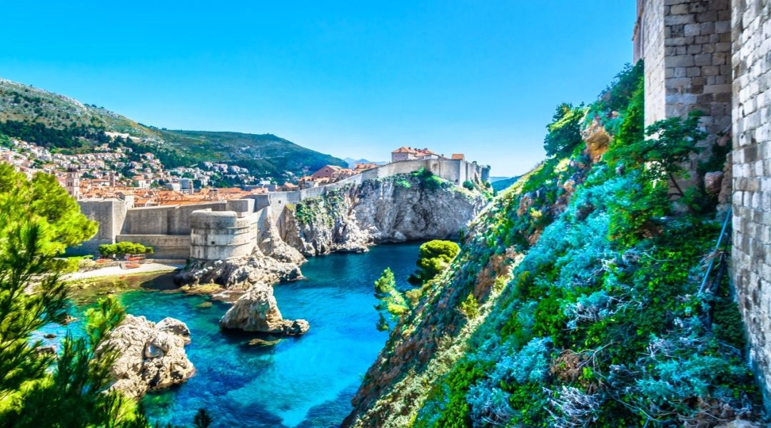 Widok na mury obronne i skaliste wybrzeże Dubrownika, błękitna woda Adriatyku i stare miasto w tle.