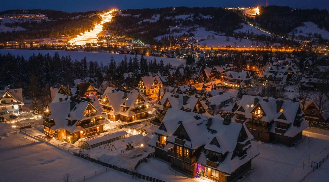 Oświetlone drewniane domy w górskiej miejscowości nocą, śnieg na dachach i stok narciarski w tle.