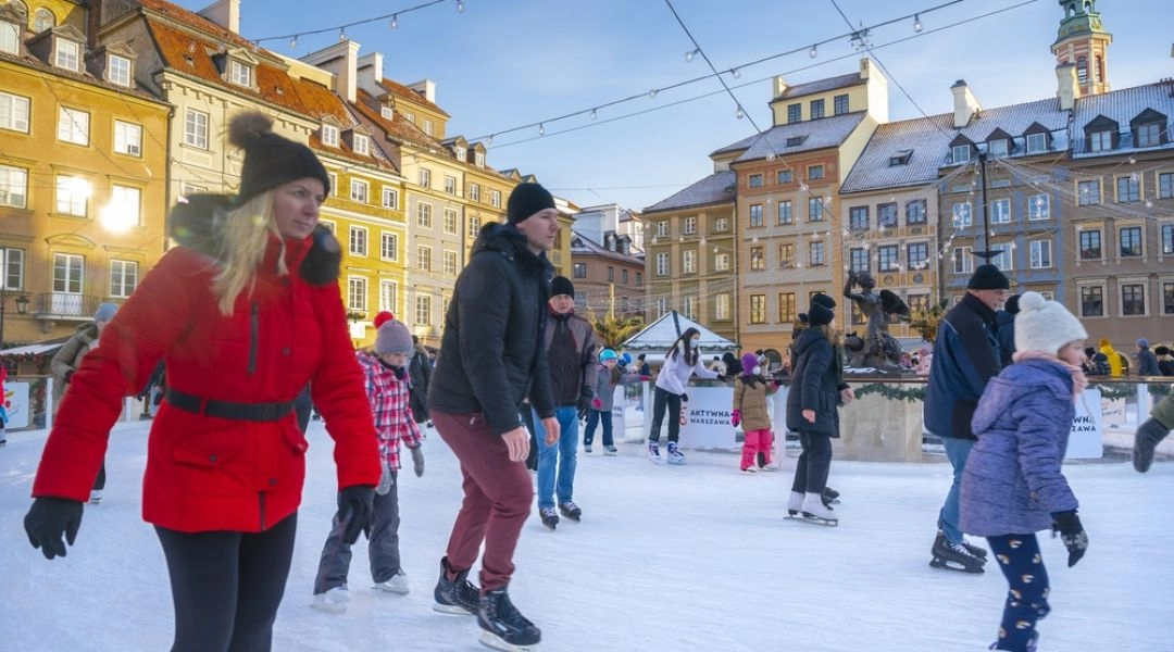 Ludzie jeżdżą na łyżwach na miejskim lodowisku w Warszawie, dzieci i dorośli na lodzie, zabudowa miejska w tle.