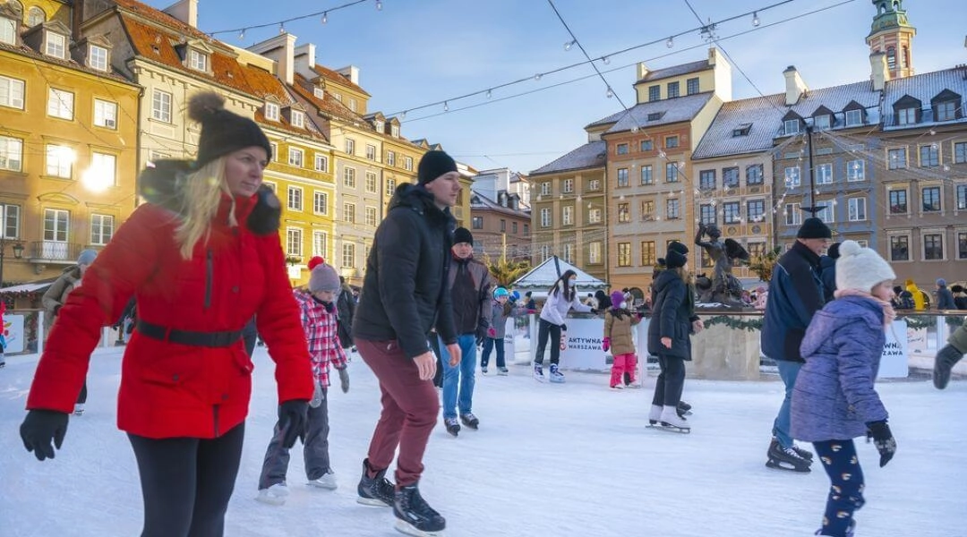 Ludzie jeżdżą na łyżwach na miejskim lodowisku w Warszawie, dzieci i dorośli na lodzie, zabudowa miejska w tle.