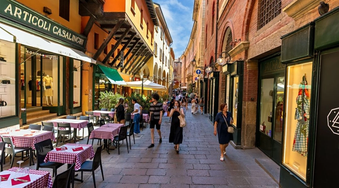 Wąska ulica w centrum Bolonii z restauracyjnymi stolikami, sklepami i spacerującymi turystami.