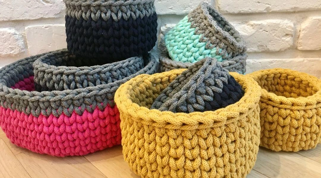 Ręcznie wykonane koszyki ze sznurka w kilku kolorach, ustawione na podłodze przy jasnej ścianie.