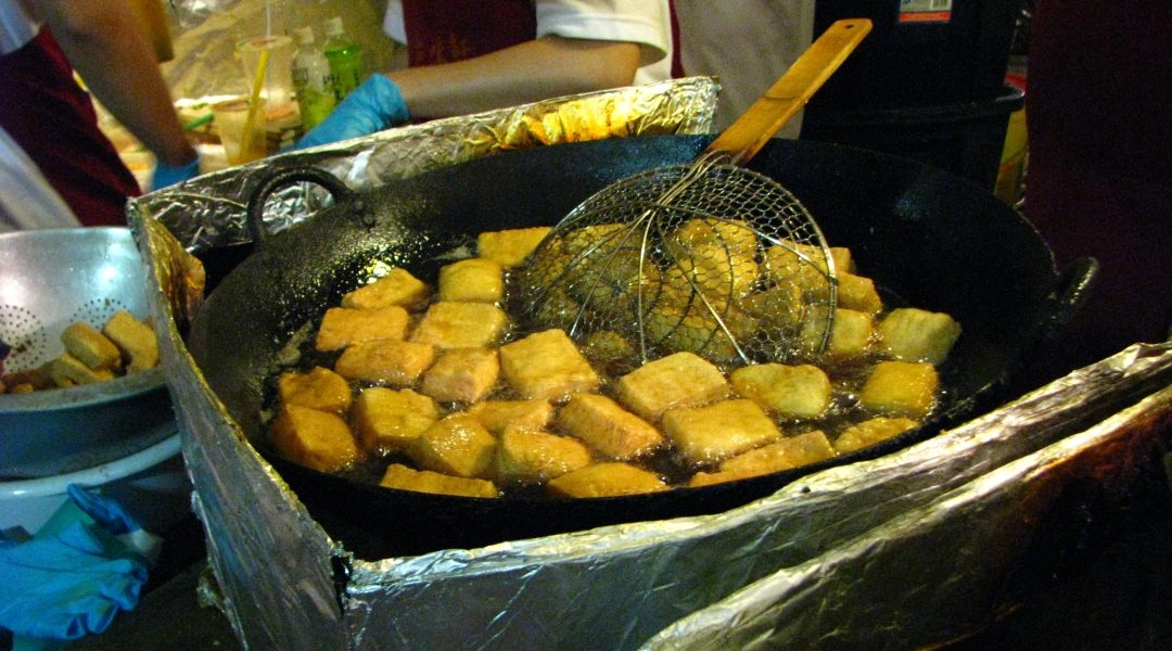 Prostokątne kawałki tofu smażące się w oleju w dużym czarnym woku z metalowym cedzakiem.