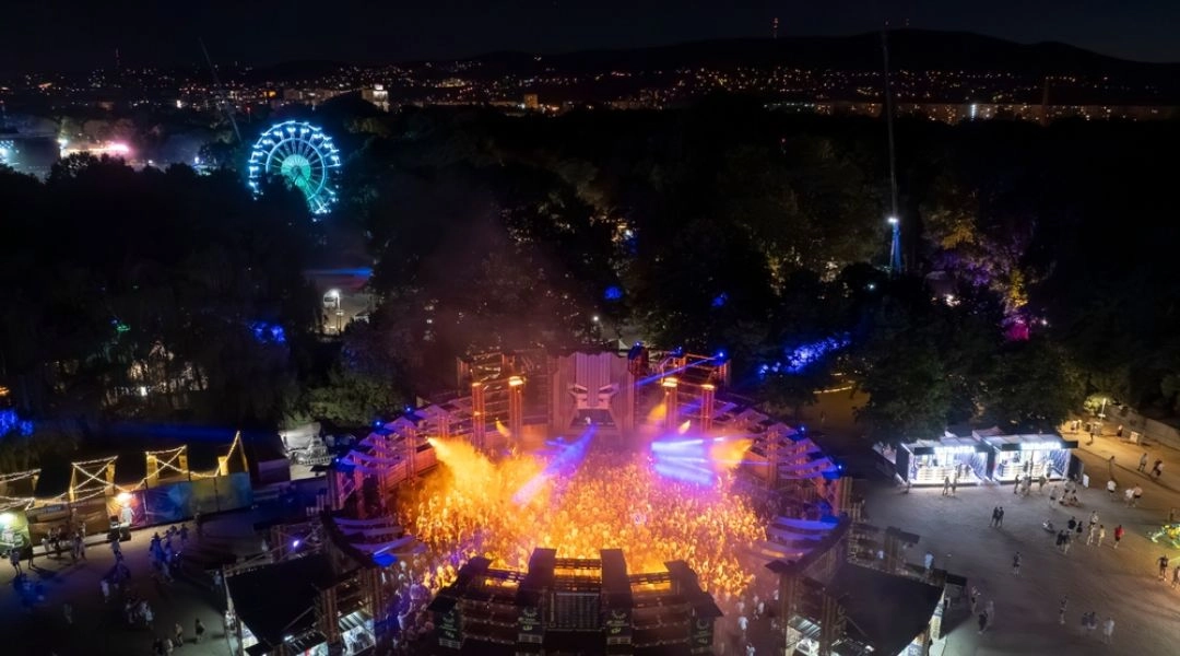 Nocny widok z góry na scenę Sziget Festival, tłum uczestników, kolorowe światła oraz podświetlone koło widokowe wśród drzew.