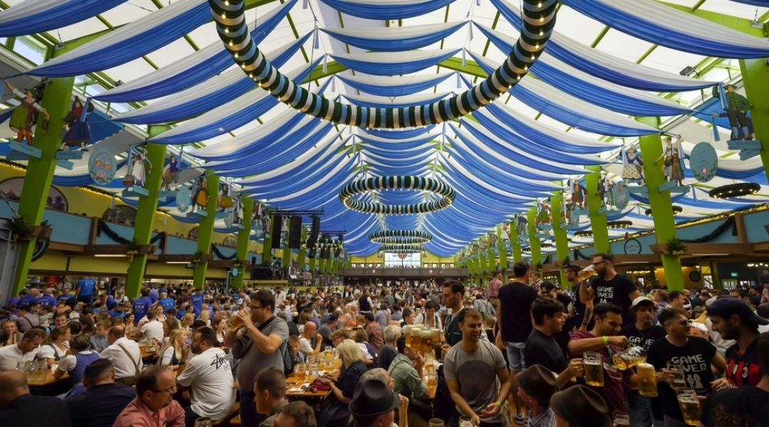 Wypełniony gośćmi namiot piwny na Oktoberfeście, z długimi stołami, zielonymi słupami i biało-niebieskimi dekoracjami pod sufitem.