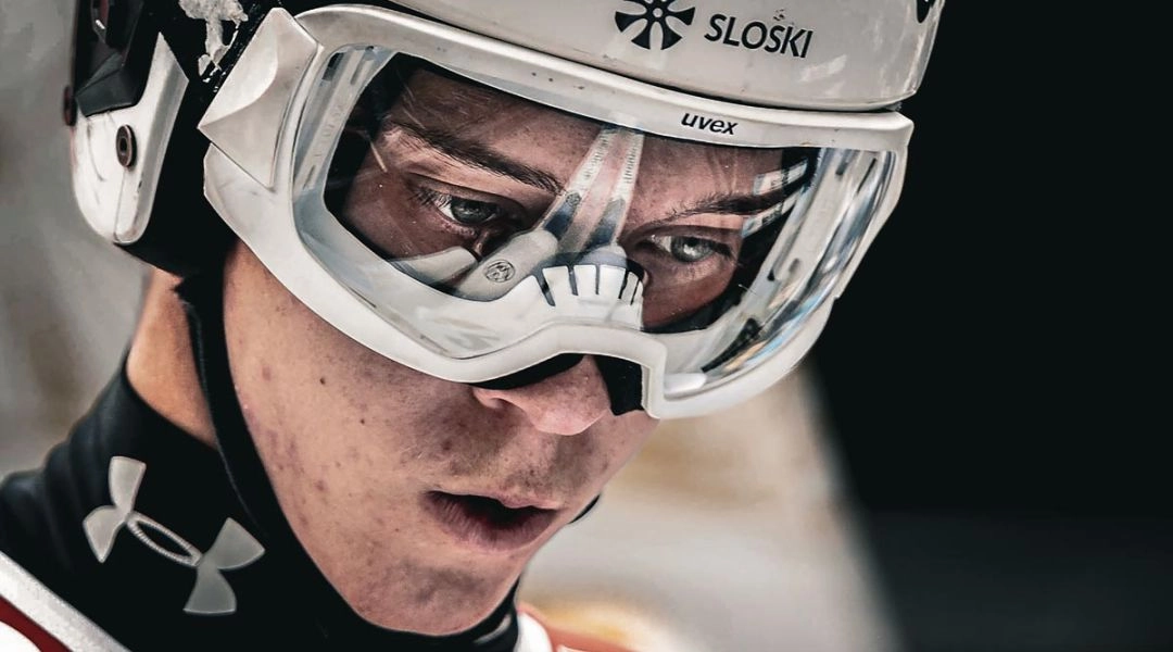 Zbliżenie na twarz skoczka narciarskiego w kasku i goglach, skupionego i przejętego tuż przed oddaniem skoku.