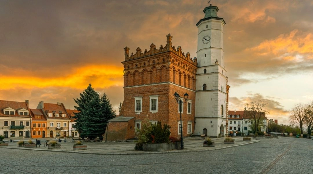 Ceglany ratusz w Sandomierzu z białą wieżą zegarową na brukowanym rynku pod niebem rozświetlonym zachodzącym słońcem.