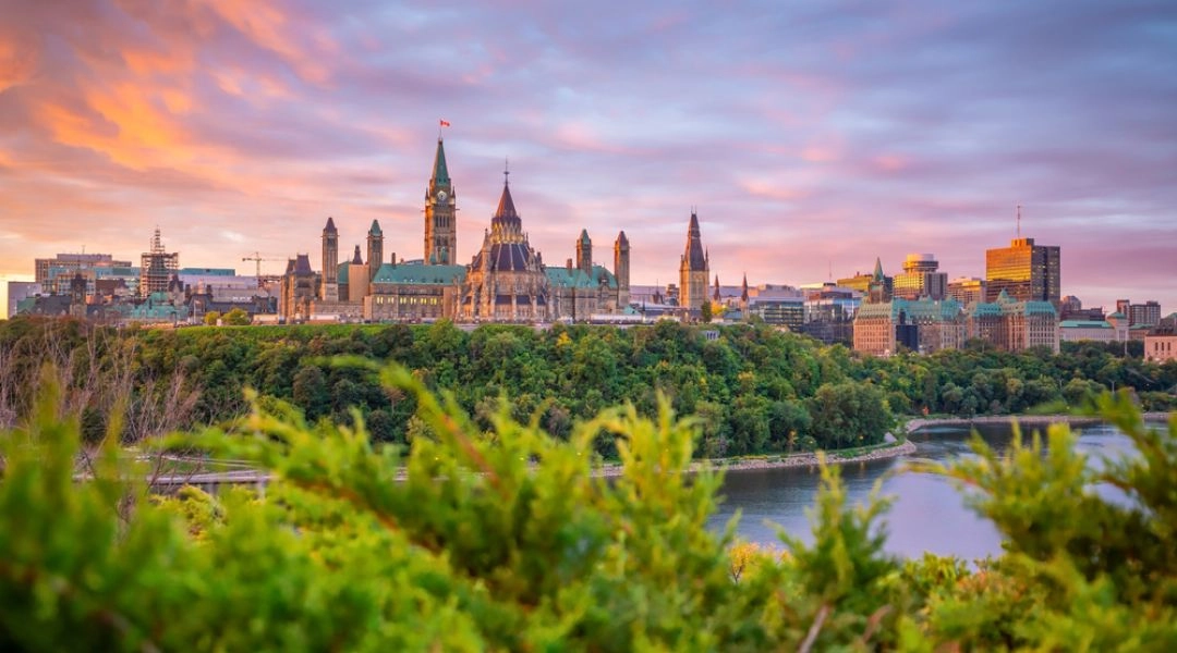 Panorama Ottawy z Parliament Hill, zielenią nad rzeką i kolorowym niebem o zachodzie słońca.