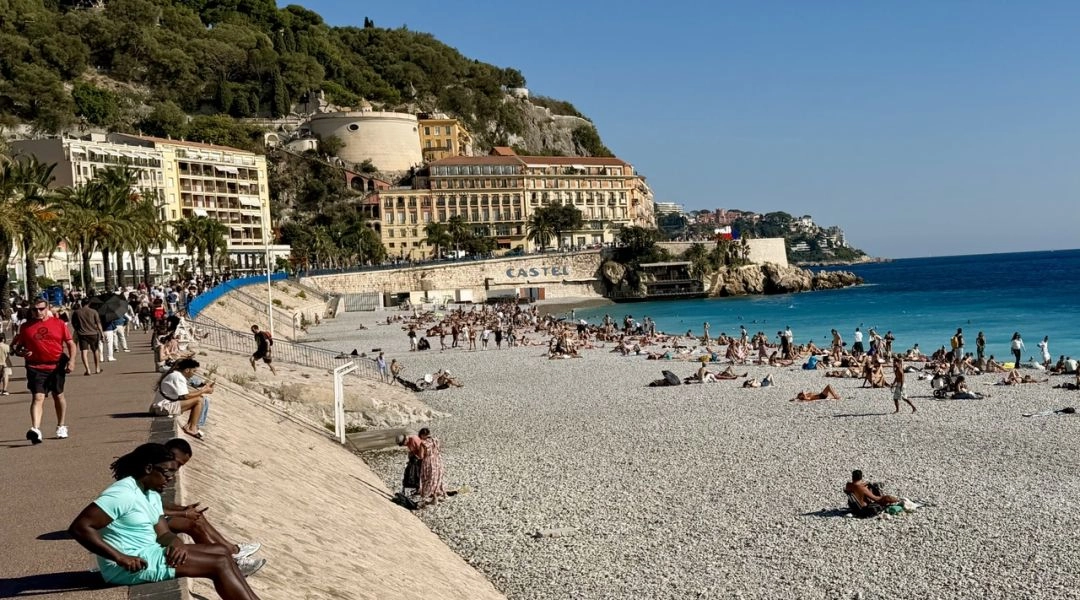 Kamienista plaża w Nicei nad turkusowym Morzem Śródziemnym, promenada z palmami i zabudowa u podnóża zielonego Wzgórza Zamkowego.