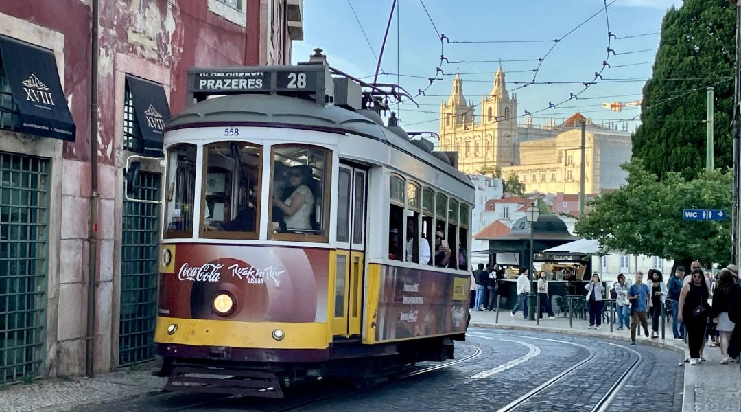 Zabytkowy tramwaj numer 28 w kremowo bordowym malowaniu jedzie po brukowanej ulicy Lizbony, a w tle widać oświetloną słońcem fasadę klasztoru São Vicente de Fora.