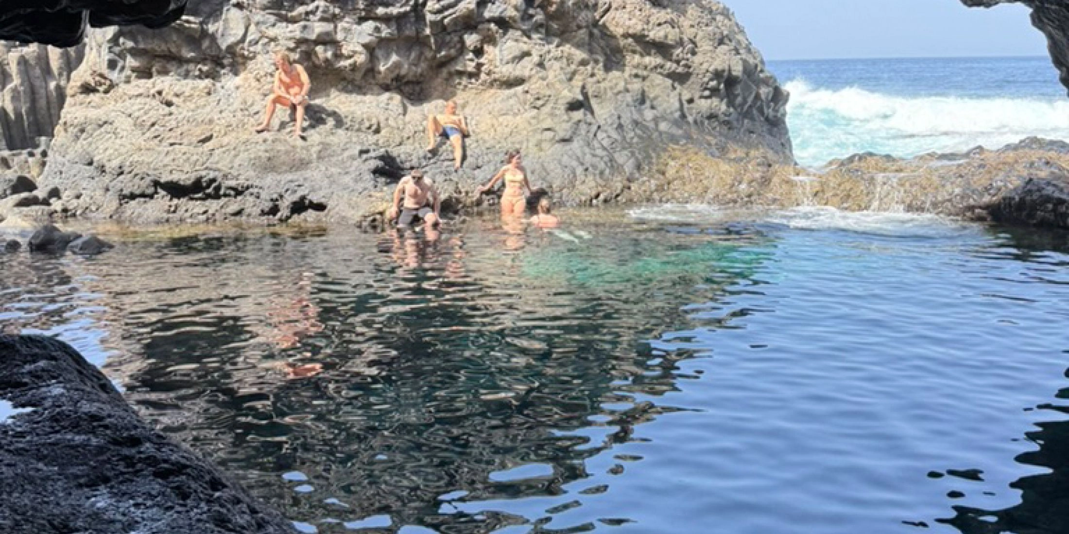 Naturalny basen lawowy na El Hierro z osobami kąpiącymi się w ciemnej skalnej grocie nad oceanem.