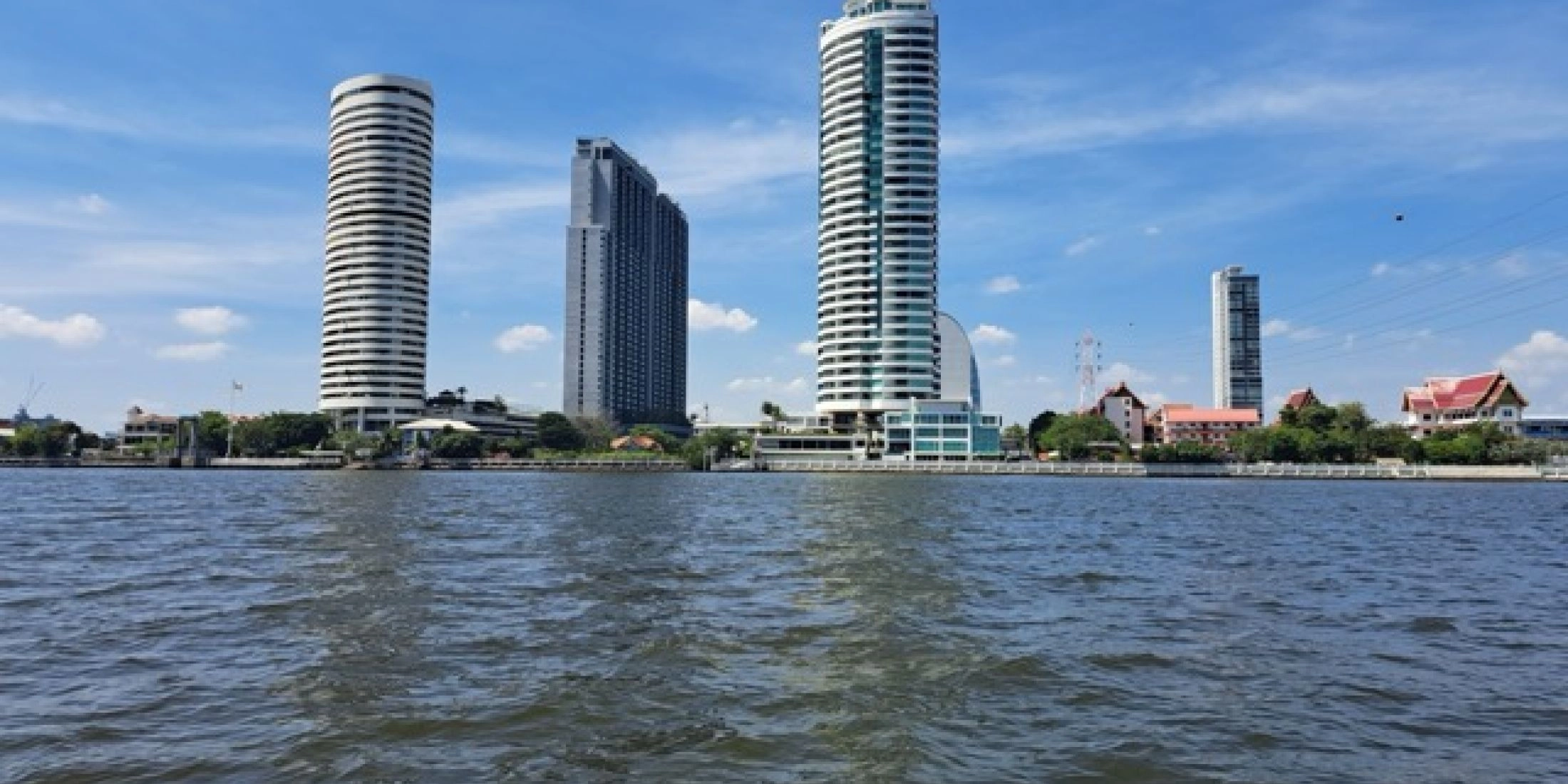 Panorama Bangkoku widziana z rzeki Chao Phraya, z nowoczesnymi wieżowcami i tradycyjnymi świątyniami w tle pod błękitnym niebem.