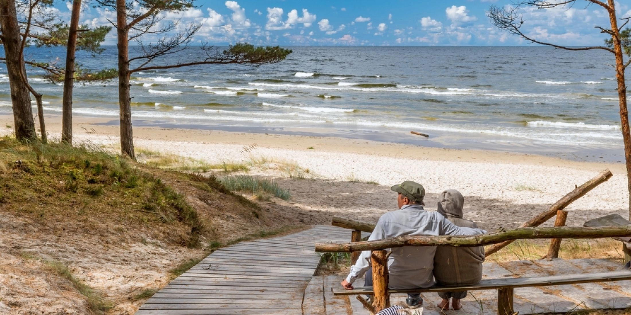 Jesienna plaża nad Bałtykiem z drewnianą ścieżką i parą siedzącą na ławce, fale i sosny nadmorskie.