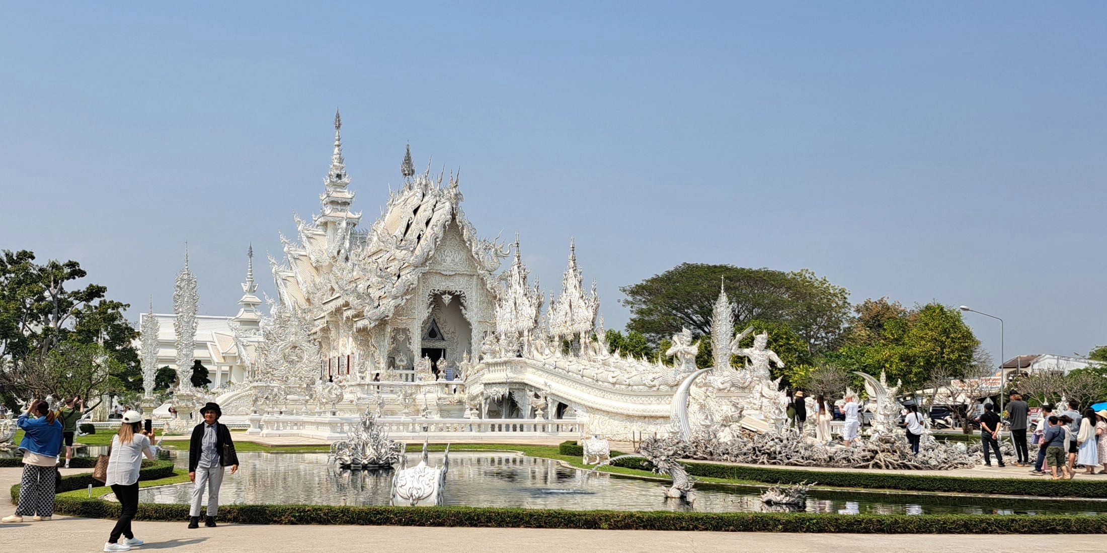 Wat Rong Khun w Chiang Rai, Biała Świątynia z rzeźbioną fasadą i mostem nad stawem, Tajlandia.