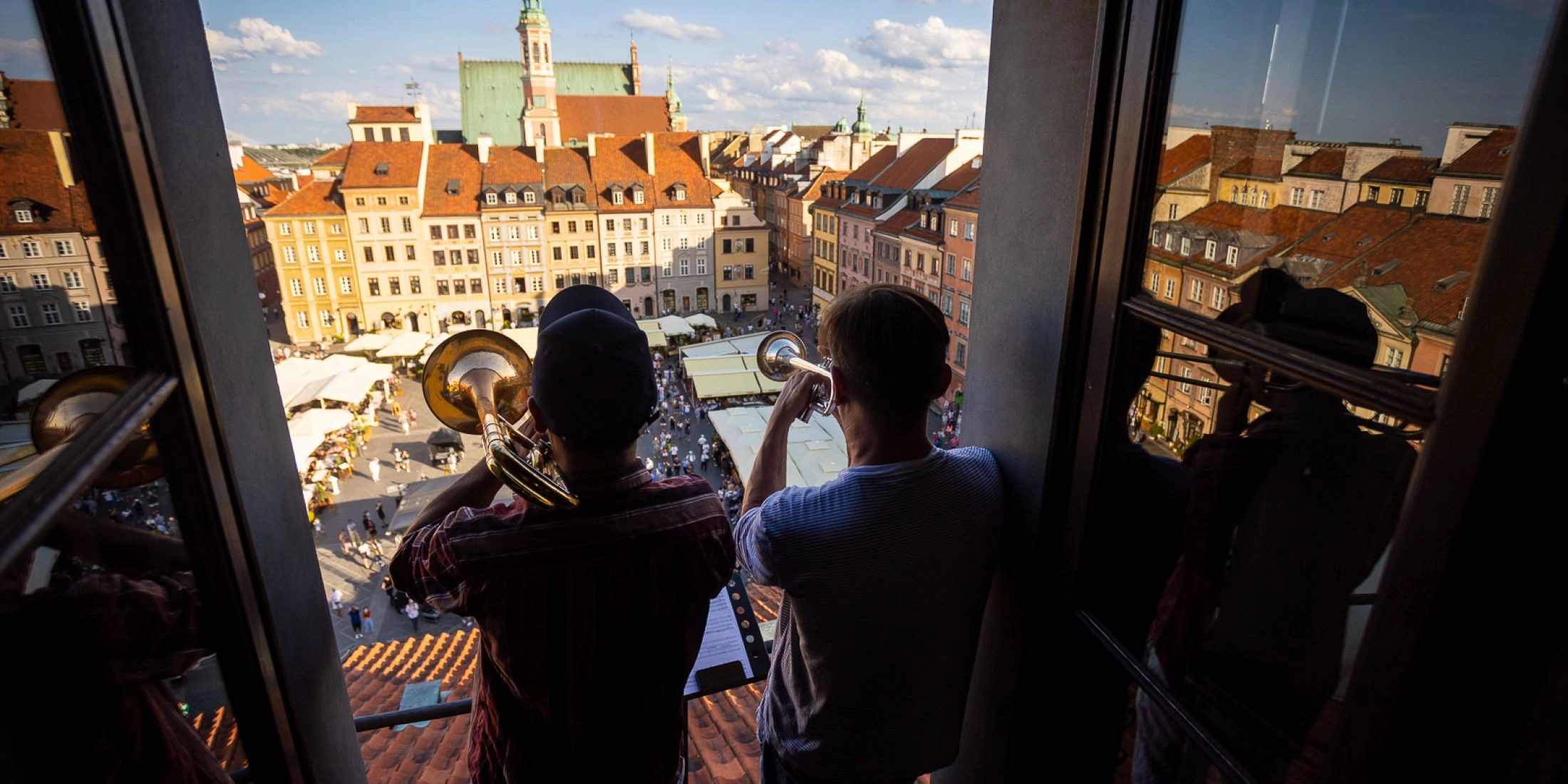 Muzycy Brass Federacji grają na instrumentach dętych w oknie kamienicy z widokiem na Rynek Starego Miasta w Warszawie.
