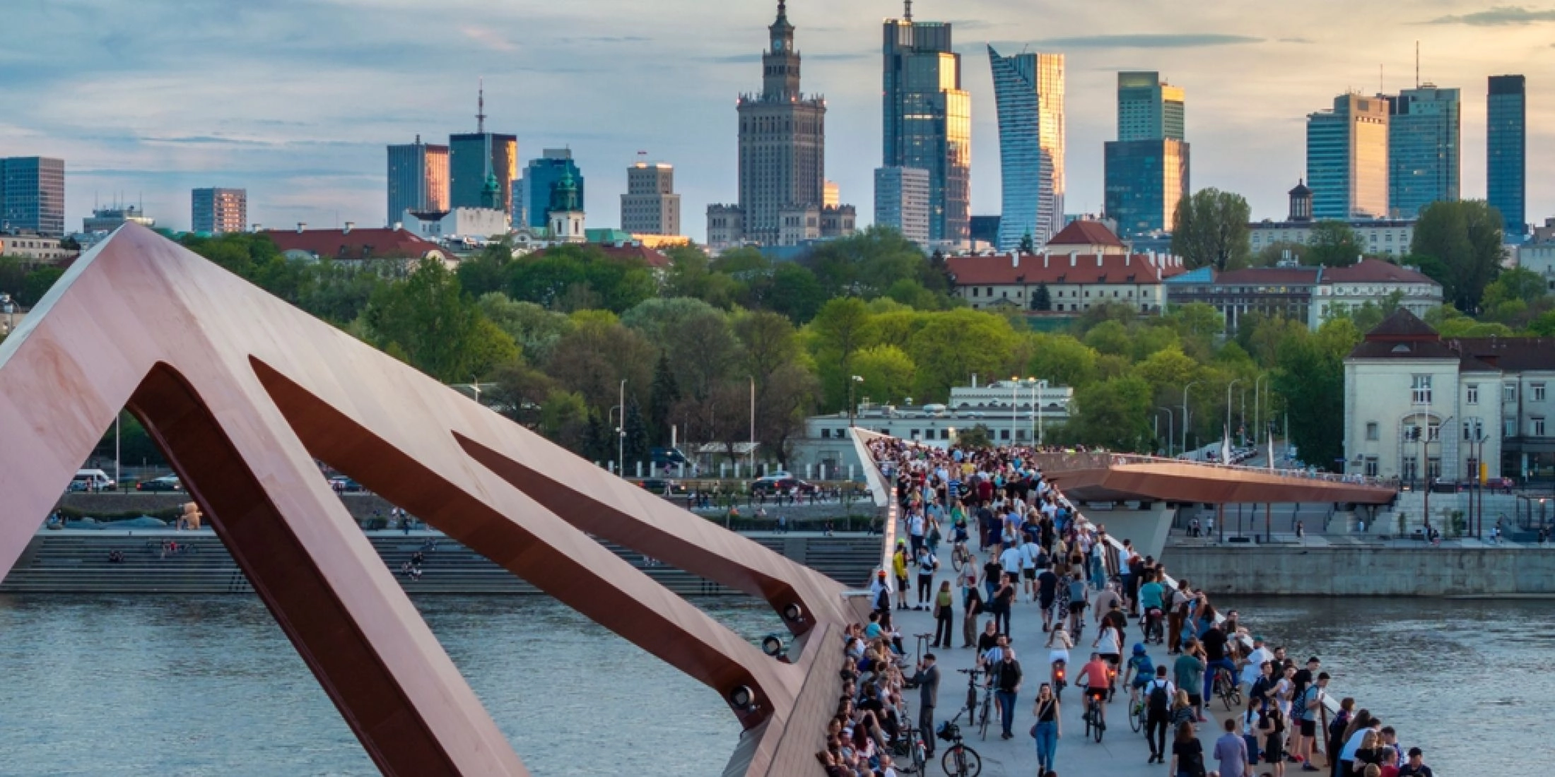 Tłum spacerowiczów i rowerzystów na kładce pieszo-rowerowej nad Wisłą w Warszawie, z panoramą centrum w tle.