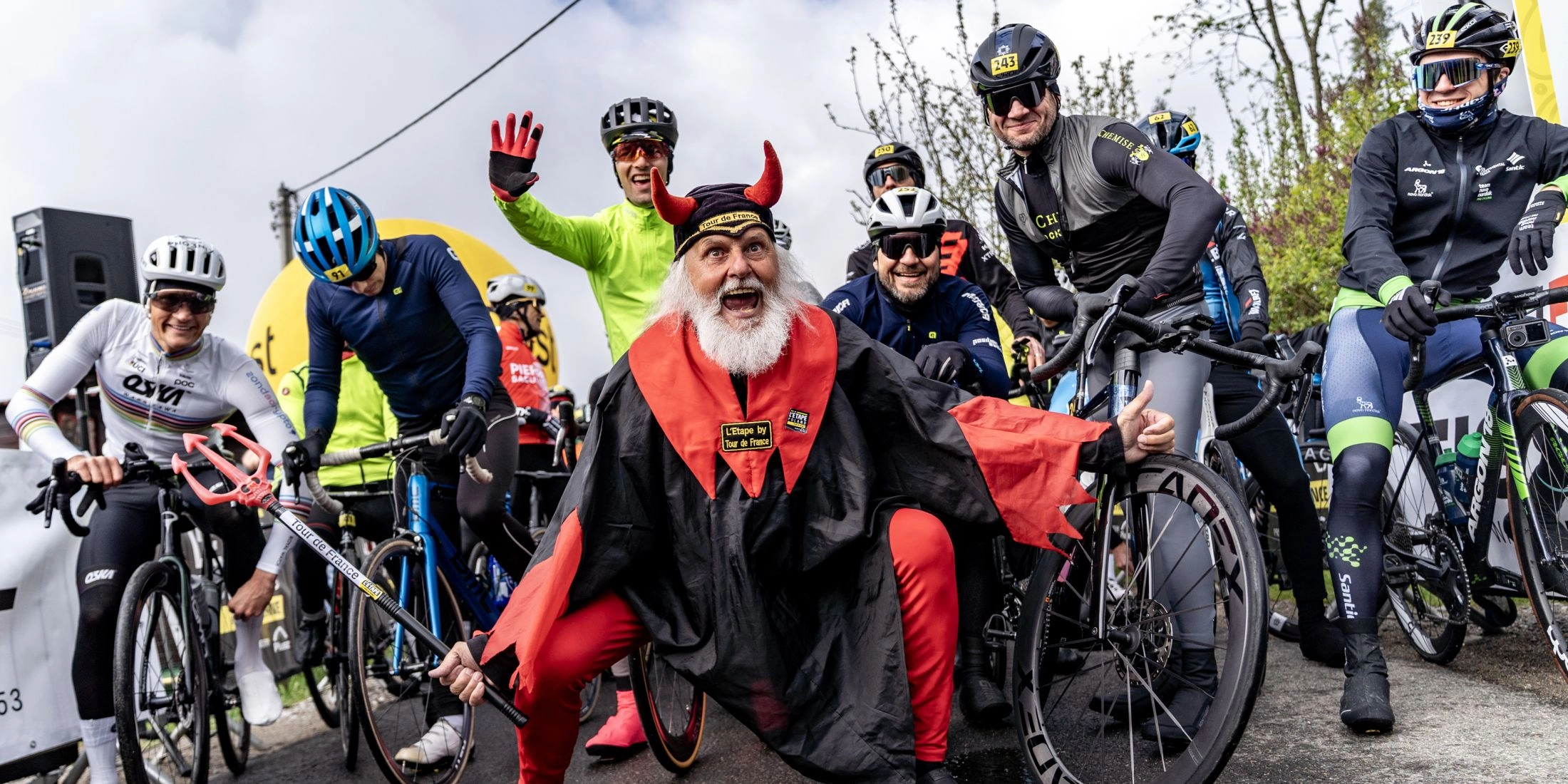 Kolarze stoją na starcie wydarzenia L’Étape Poland by Tour de France, obok nich maskotka Tour de France pozuje do zdjęcia.