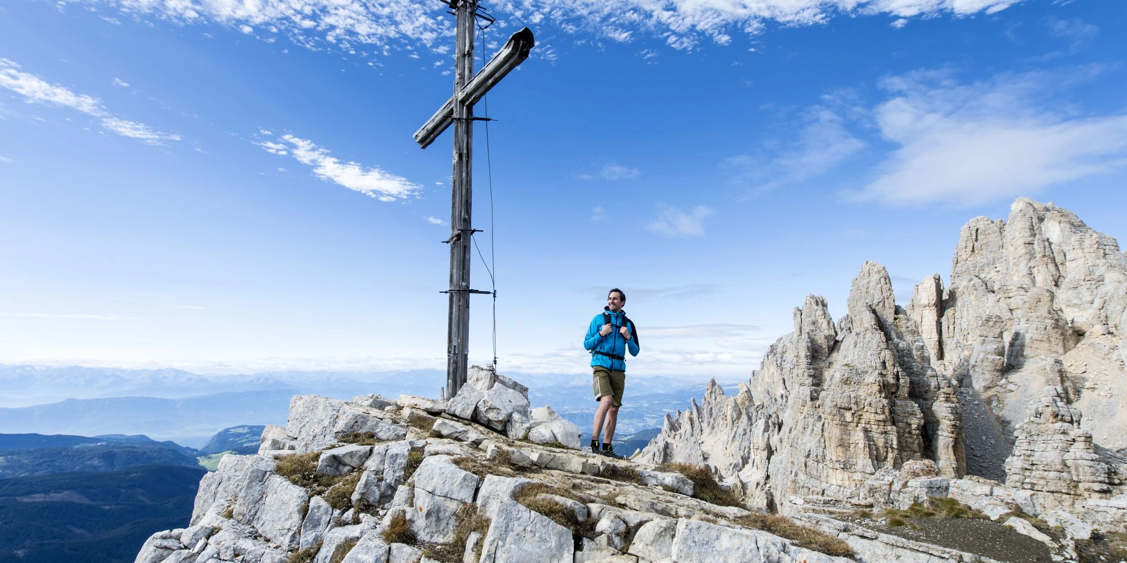 Turysta stojący obok drewnianego krzyża na skalistym szczycie, w tle jasne turnie Dolomitów i rozległa panorama gór.