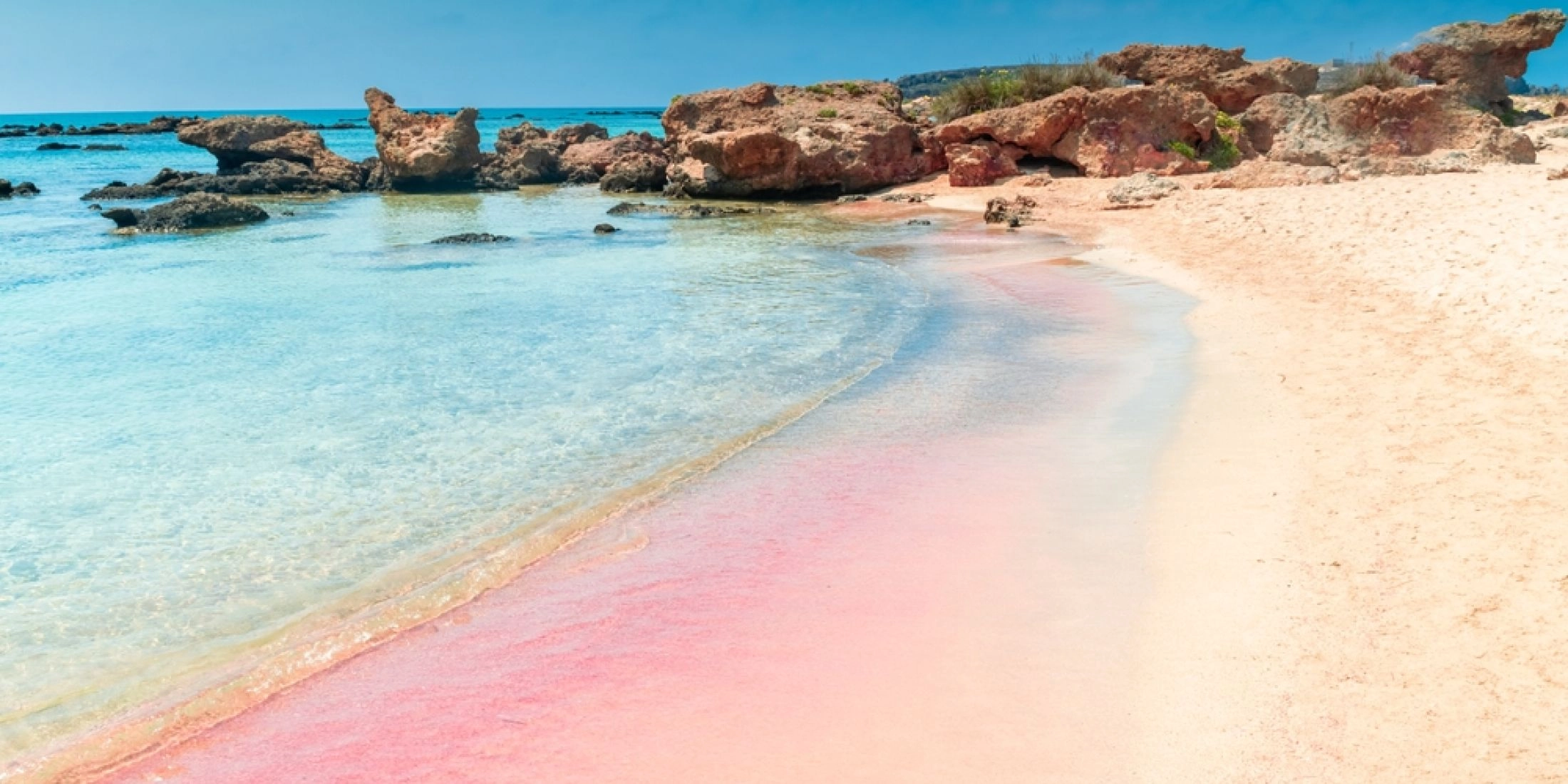 Plaża Elafonisi na Krecie z jasną wodą, różowym piaskiem i niskimi skałami przy brzegu.