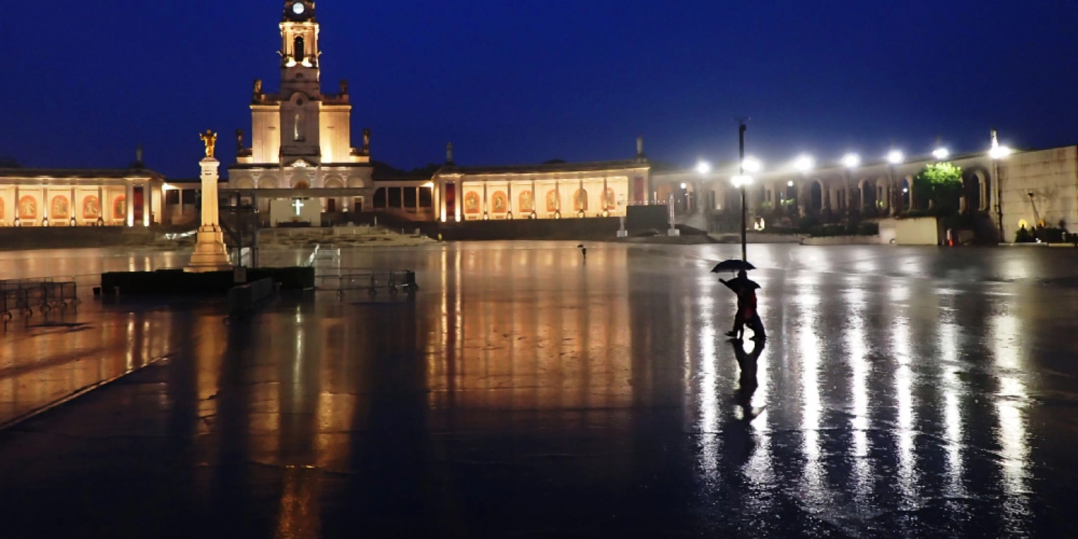 Plac sanktuarium w Fatimie nocą, z oświetloną bazyliką i postacią z parasolem odbijającą się w mokrej nawierzchni.