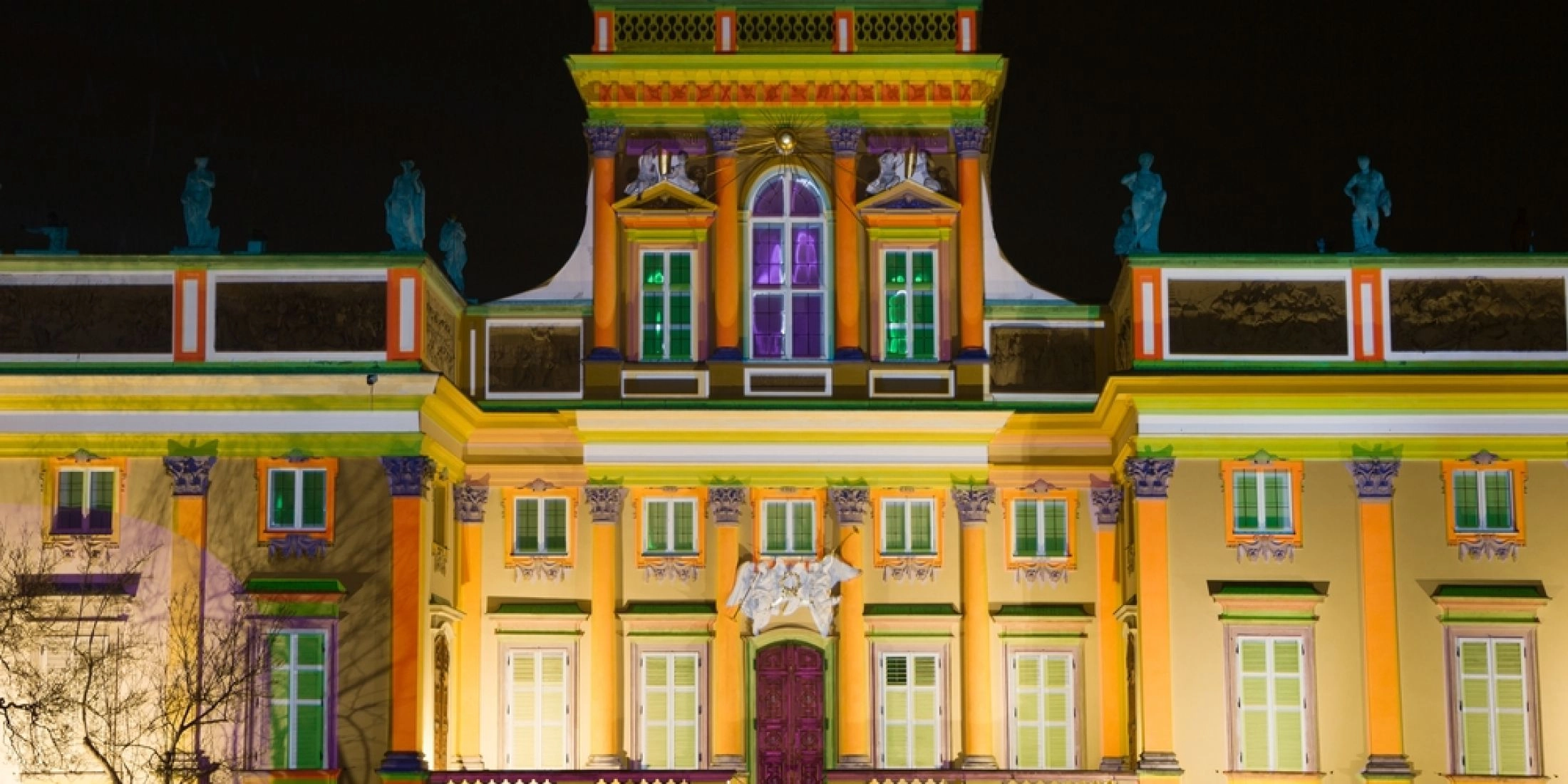 Fasada Pałacu w Wilanowie nocą, oświetlona kolorowymi iluminacjami podkreślającymi kolumny, okna i zdobienia budynku.