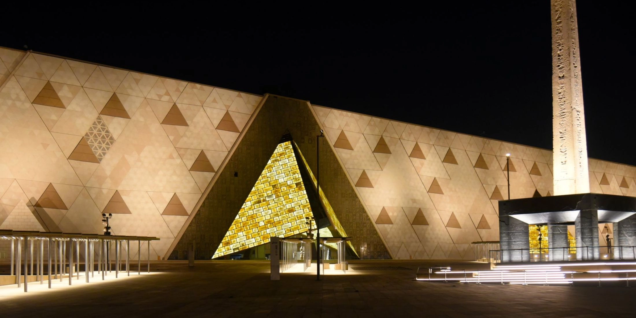 Zewnętrzna elewacja muzeum w kształcie trójkąta, iluminowana po zmroku w barwach złota i piaskowca.