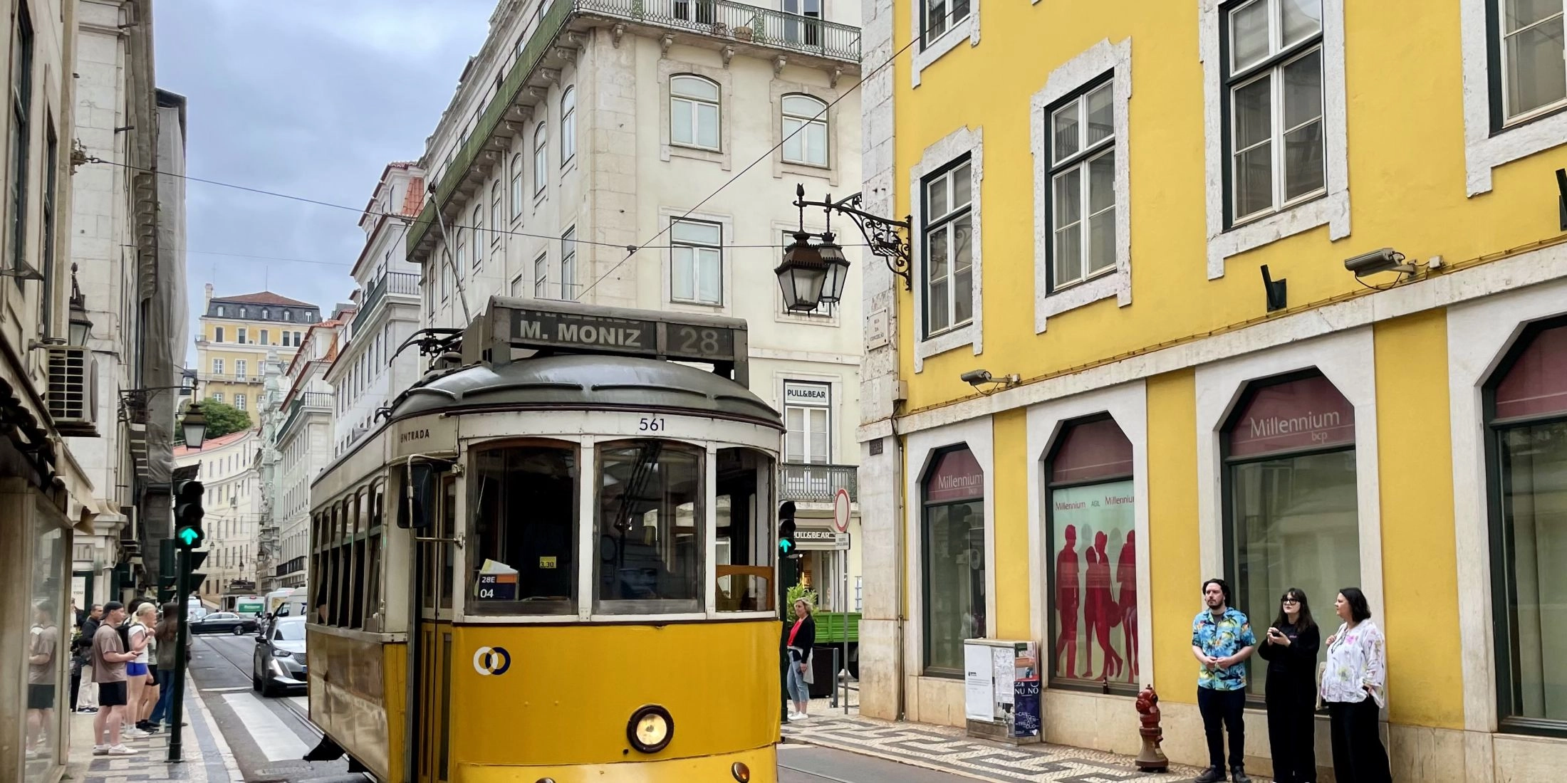 Żółty zabytkowy tramwaj numer 28 jedzie wąską ulicą Lizbony między kamienicami i kolorowymi fasadami.