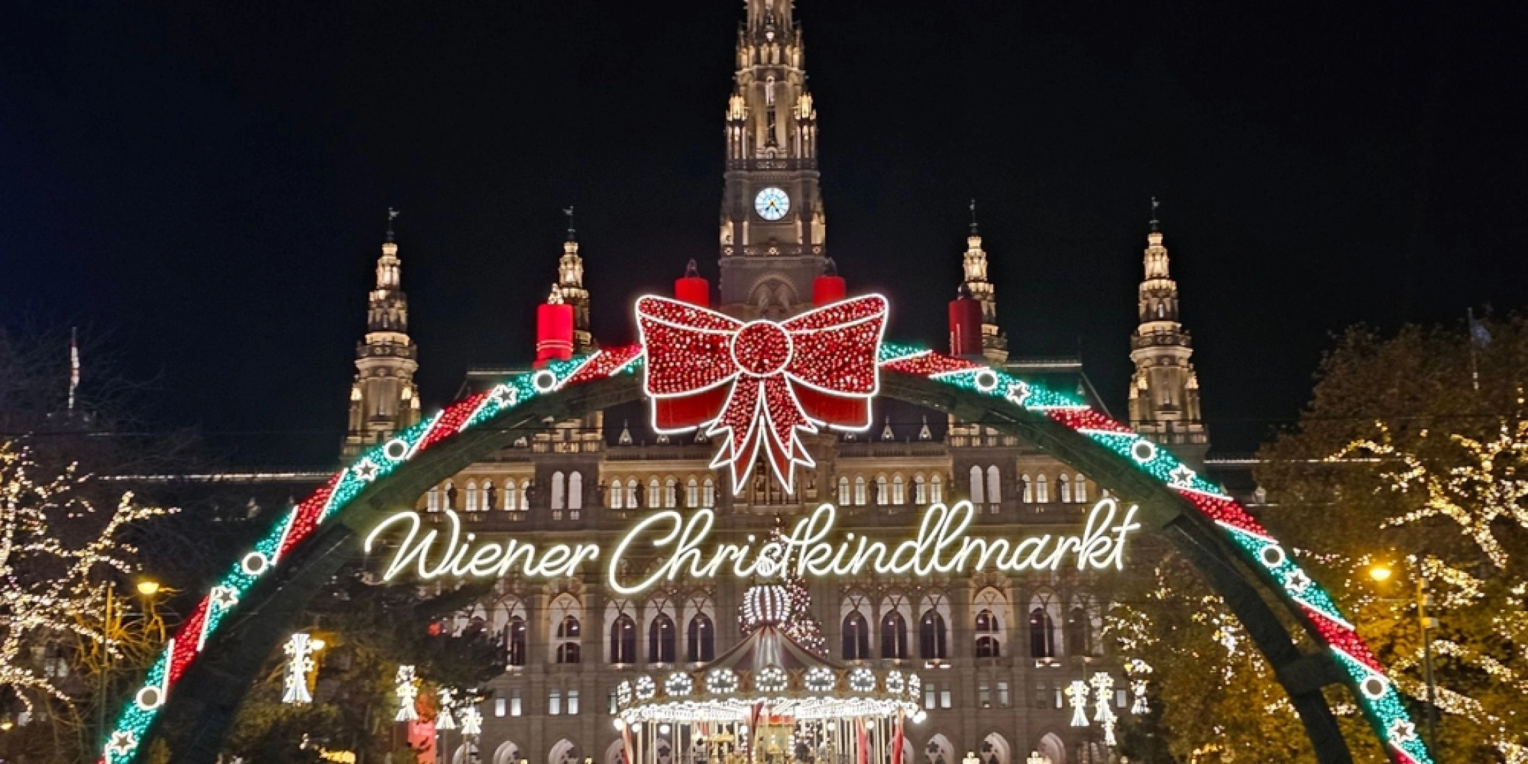 Plac przed wiedeńskim ratuszem z lodowiskiem i iluminacjami, świąteczne stoiska i odwiedzający po zmroku.