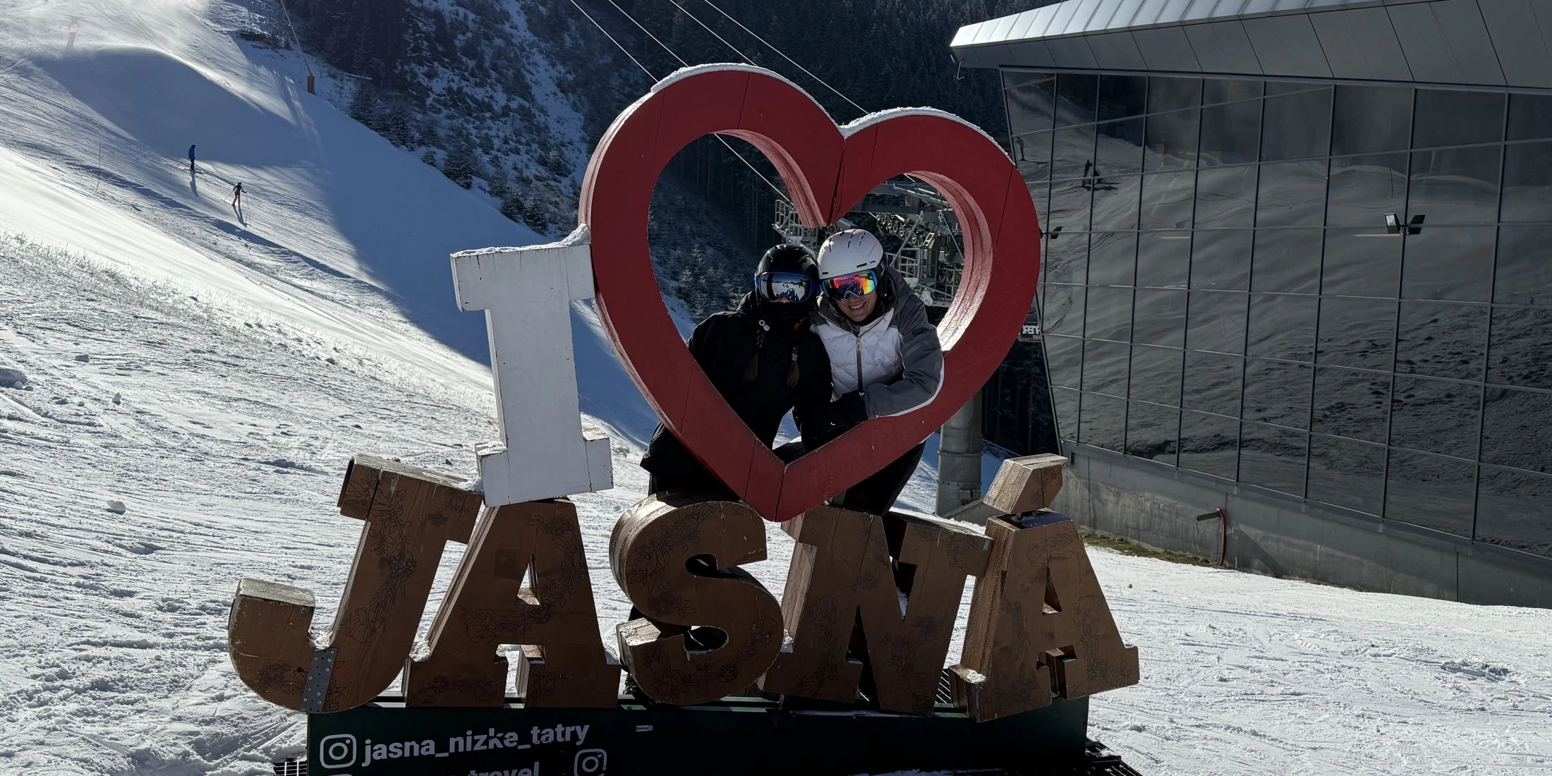 Dwoje narciarzy w kaskach i goglach pozuje w dekoracji „I love Jasná” przy stoku narciarskim i nowoczesnej stacji kolei w Jasnej w Niskich Tatrach.