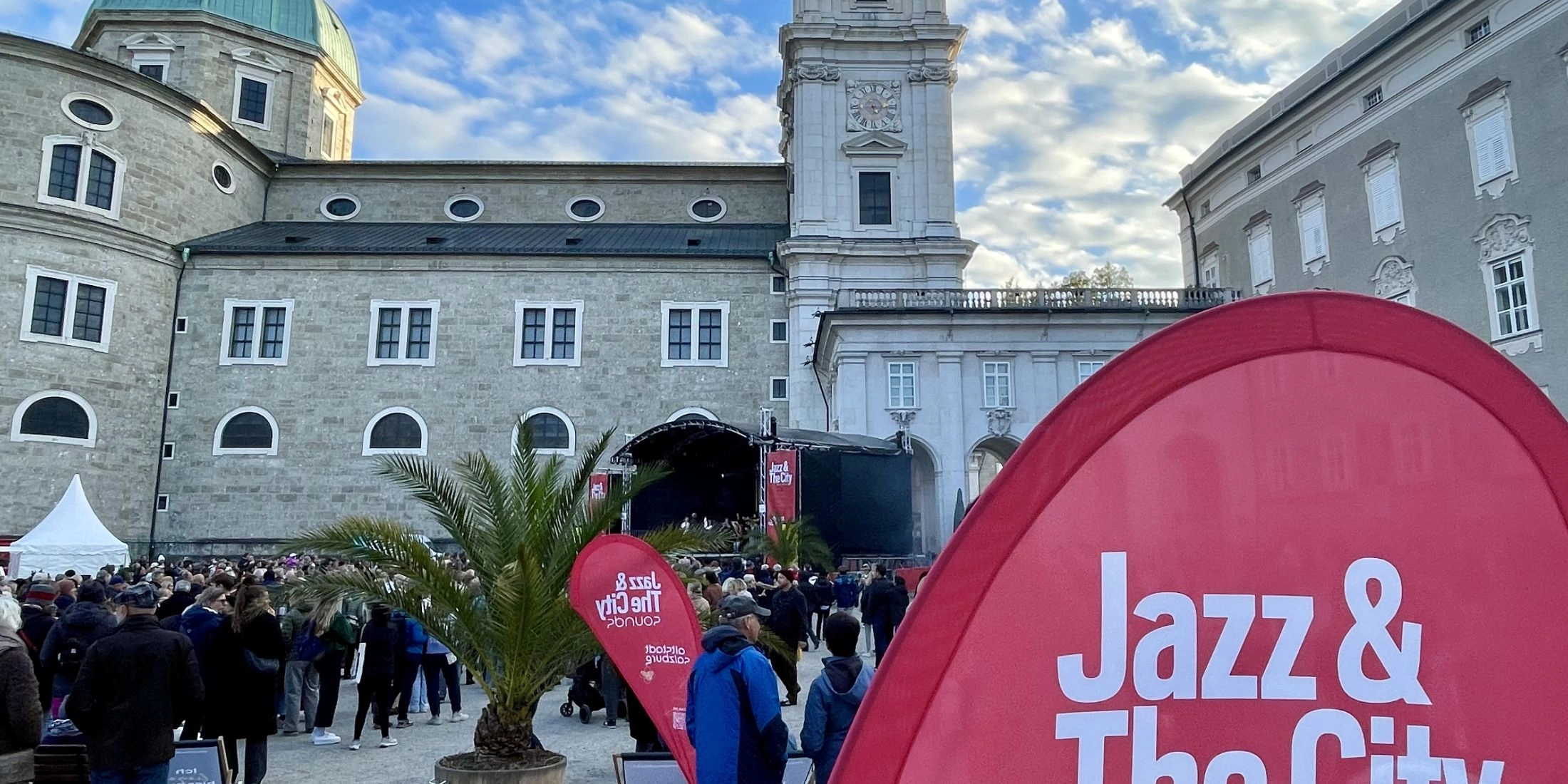 Tłum ludzi na placu Residenzplatz w Salzburgu podczas festiwalu Jazz&TheCity, w tle katedra i scena koncertowa.