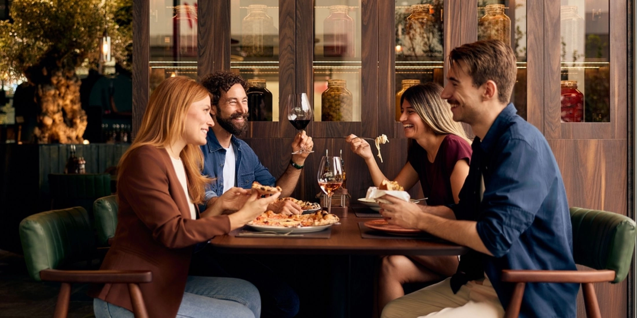 Cztery osoby siedzą przy stole w restauracji, jedzą i rozmawiają w przytulnym wnętrzu z drewnianą zabudową i półkami z przetworami w tle.