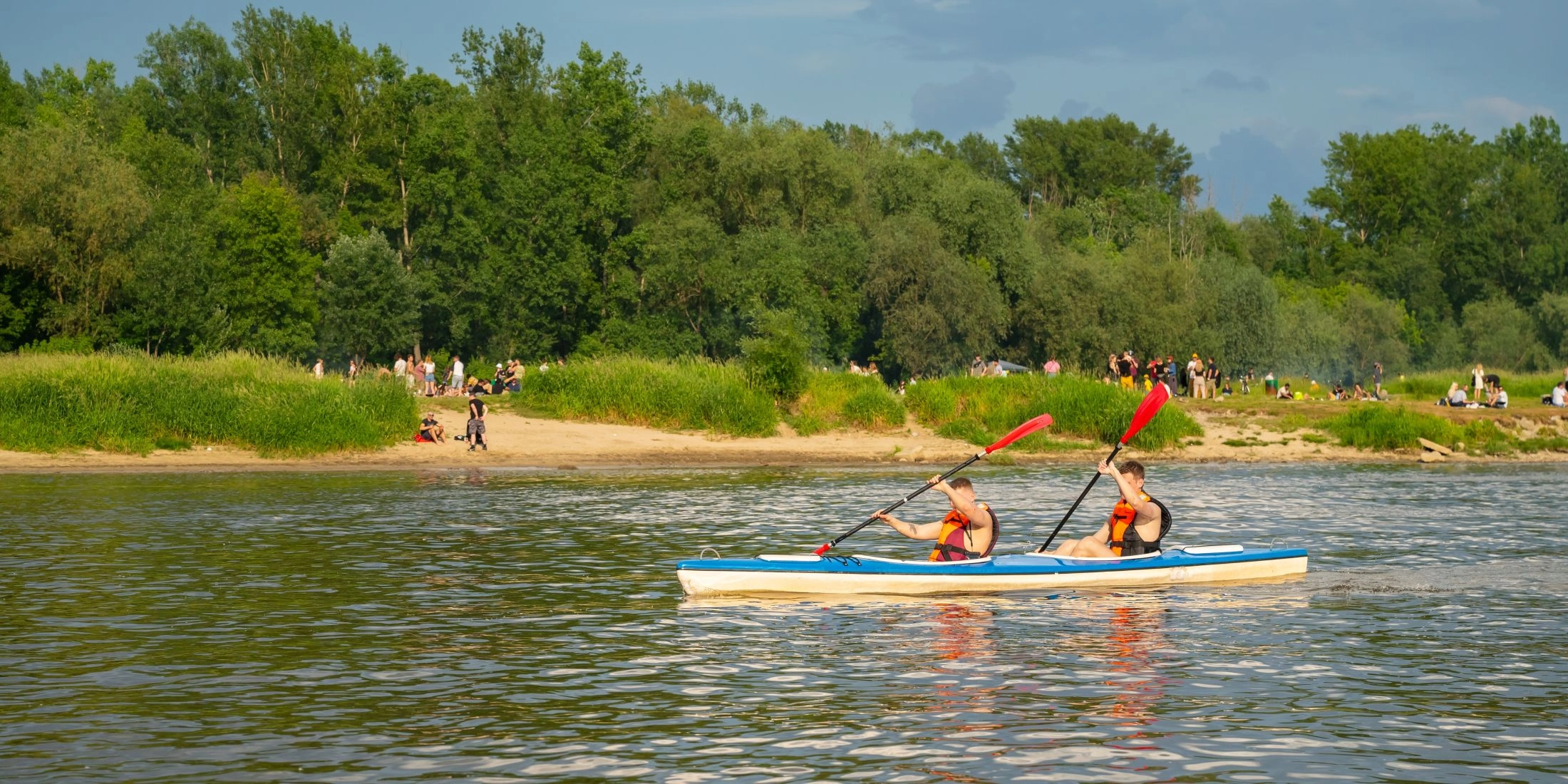 Dwie osoby w kamizelkach ratunkowych płyną dwuosobowym kajakiem po Wiśle, w tle piaszczysta plaża i zielony brzeg.