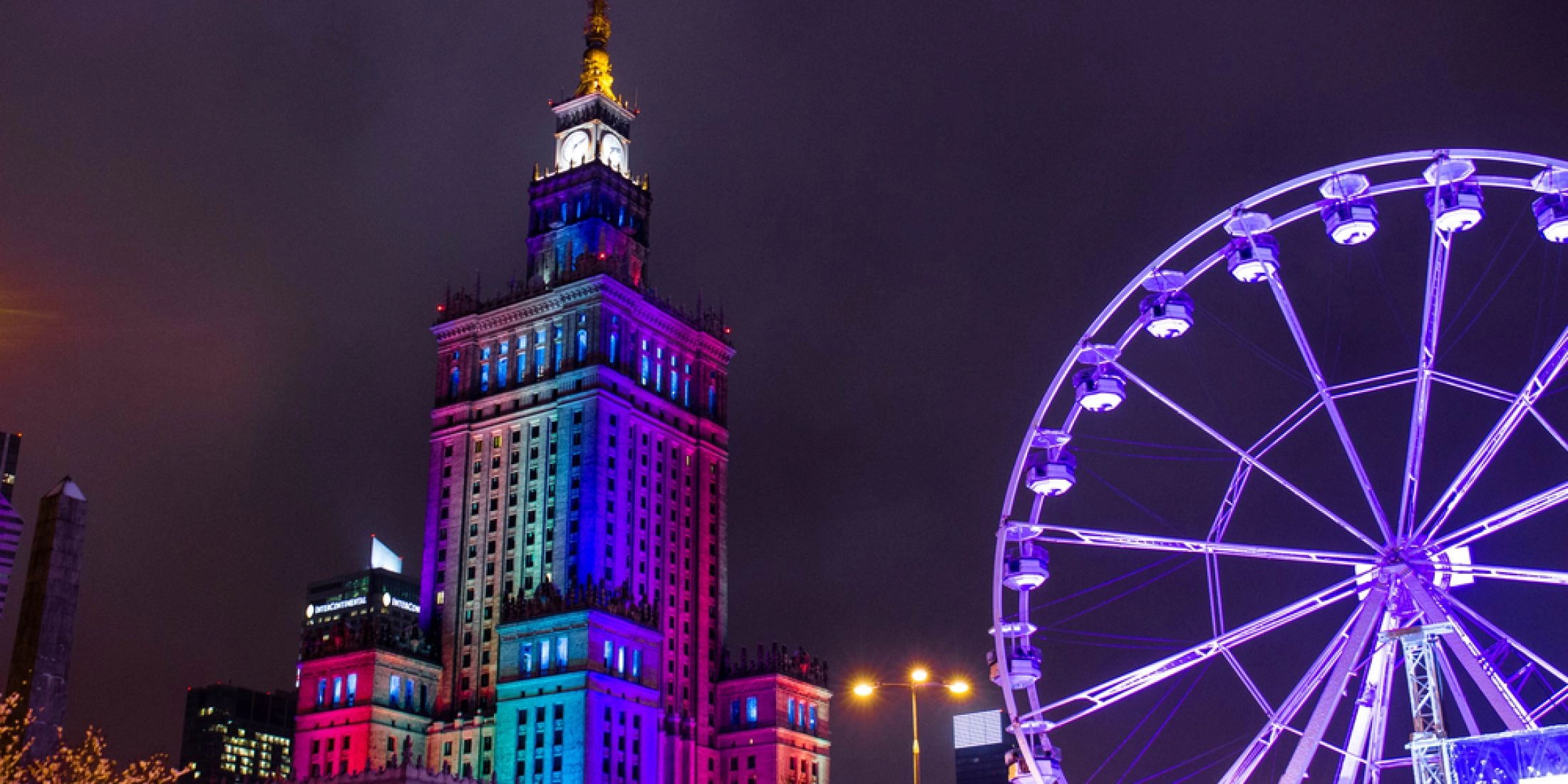 Nocne ujęcie Pałacu Kultury w kolorowych światłach z dużym diabelskim młynem na pierwszym planie.