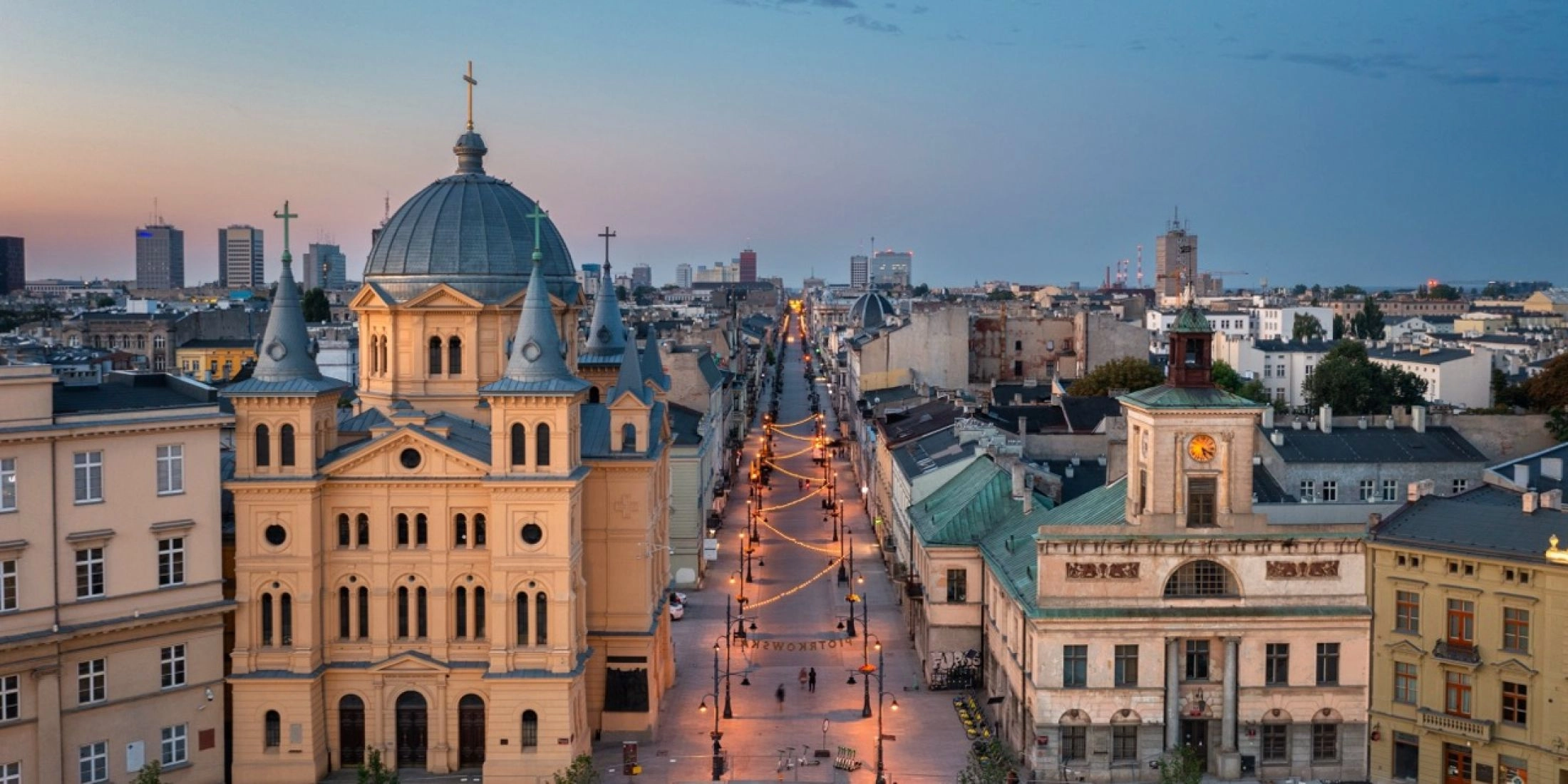 Widok z lotu ptaka na Plac Wolności i początek ulicy Piotrkowskiej w Łodzi o zmierzchu. Po lewej stronie widać charakterystyczny budynek kościoła Zesłania Ducha Świętego z kopułą, a po prawej budynek z wieżyczką zegarową. Ulica Piotrkowska jest oświetlona